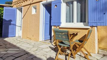 A stone-paved courtyard with a wooden table and chairs, a window with blue shutters, and a door with a blue shutter.