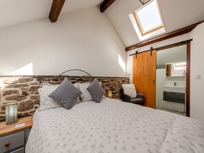 A bedroom with a stone wall, a bed with patterned bedding, a nightstand, a bedside lamp, a chair, and a wooden door leading to a bathroom.