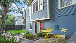 Une maison à l’extérieur bleu, avec une porte rouge et une petite terrasse dotée de chaises jaunes et d’une table.