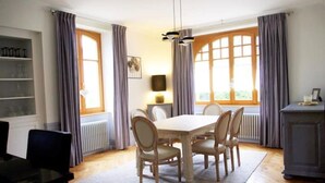 Een eetruimte met een houten tafel en stoelen, een kroonluchter en uitzicht door grote ramen.