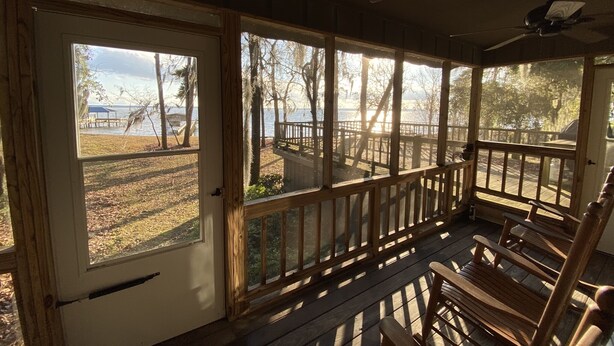 Une véranda couverte avec des balustrades en bois et un ventilateur de plafond, donnant sur un quai et des arbres.