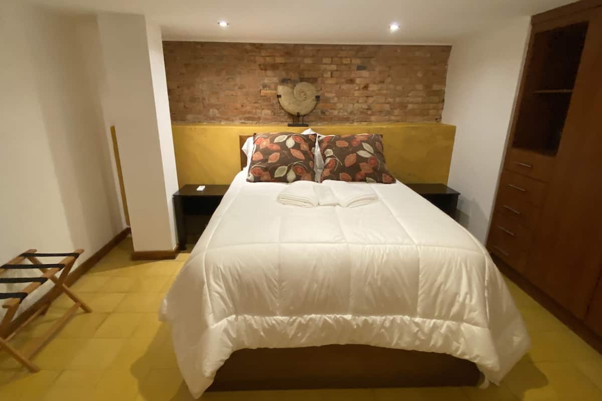A bedroom with a brick wall, a bed with white bedding, a nightstand with a fan, and a wooden wardrobe.