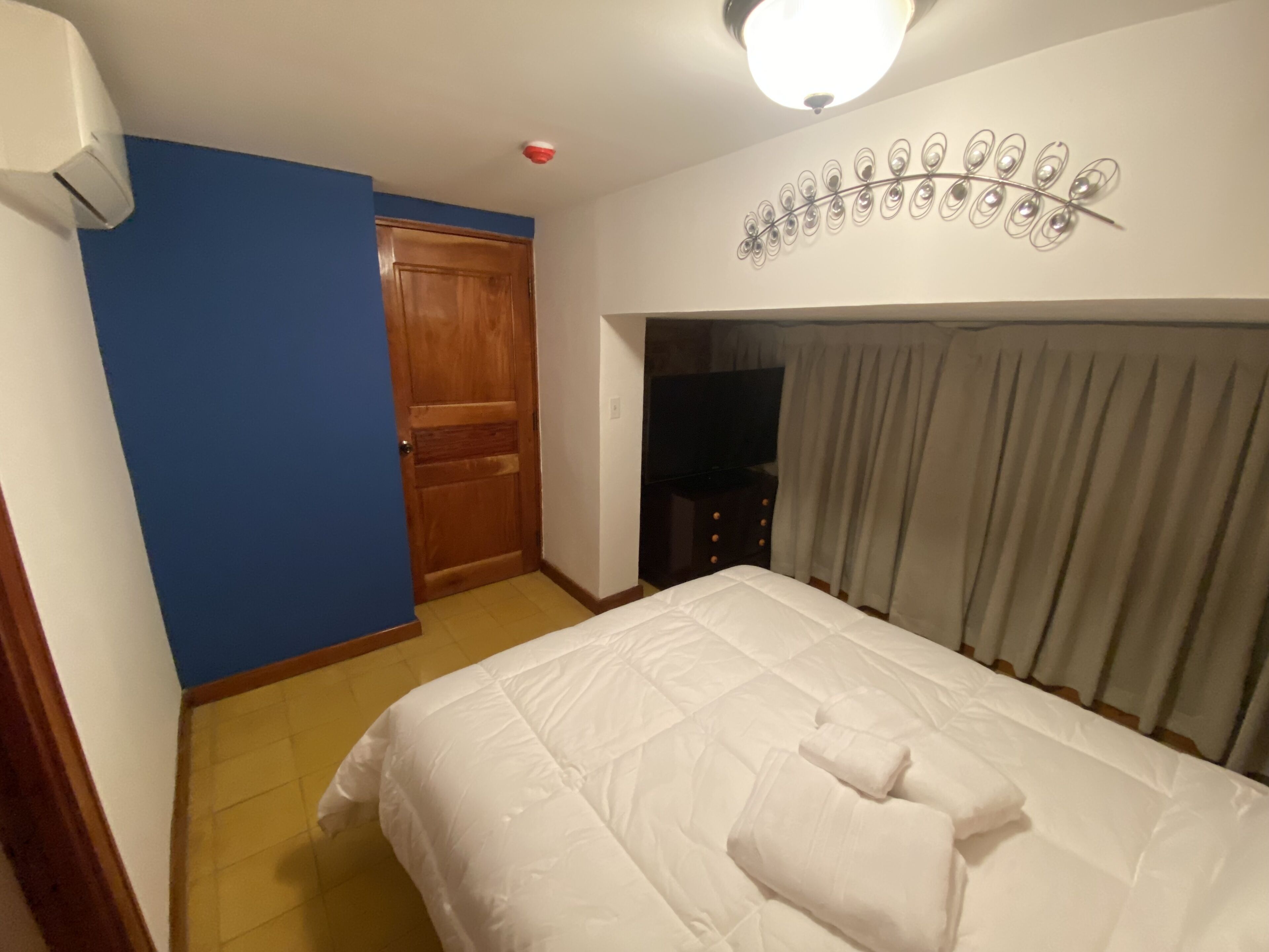 A hotel room with a bed, a wooden door, a wall-mounted air conditioner, and a ceiling light fixture.