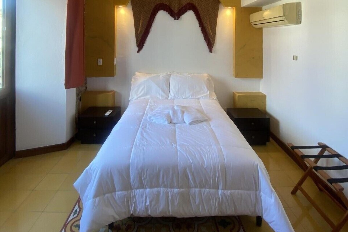 A hotel room with a bed, a chandelier, an air conditioning unit, and a patterned rug.