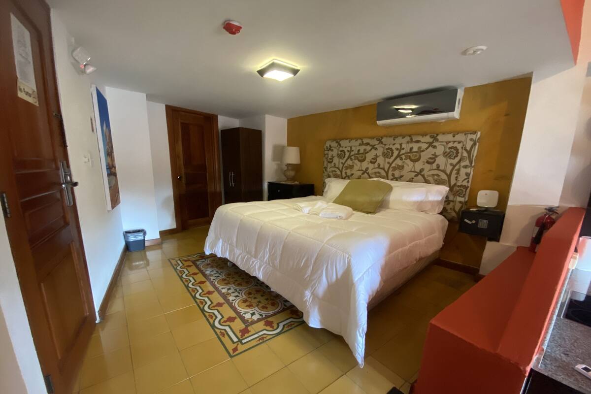 A hotel room with a bed, a nightstand, a wall-mounted air conditioner, a patterned rug, and a wooden door.