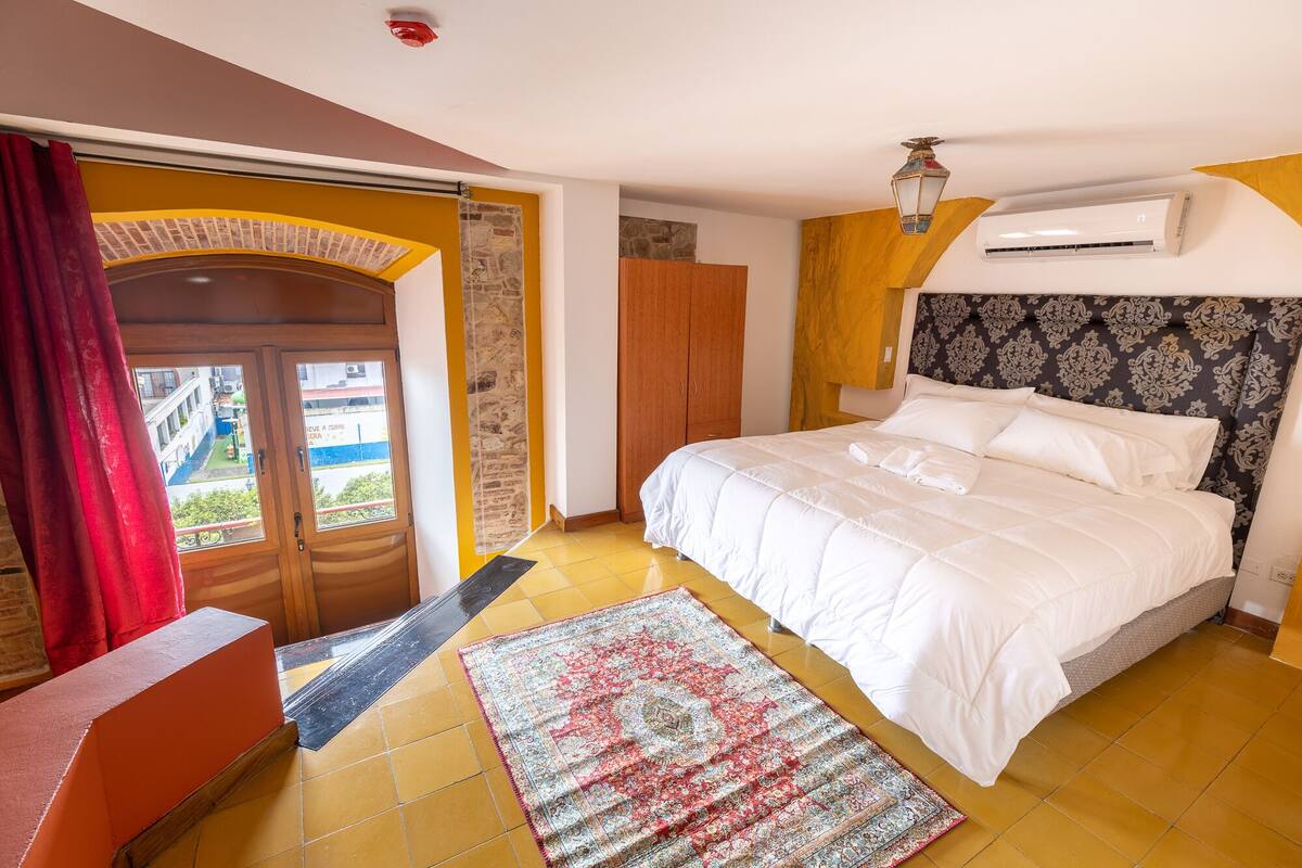 A hotel room with a bed, a view through a door, a patterned rug, and an air conditioning unit.