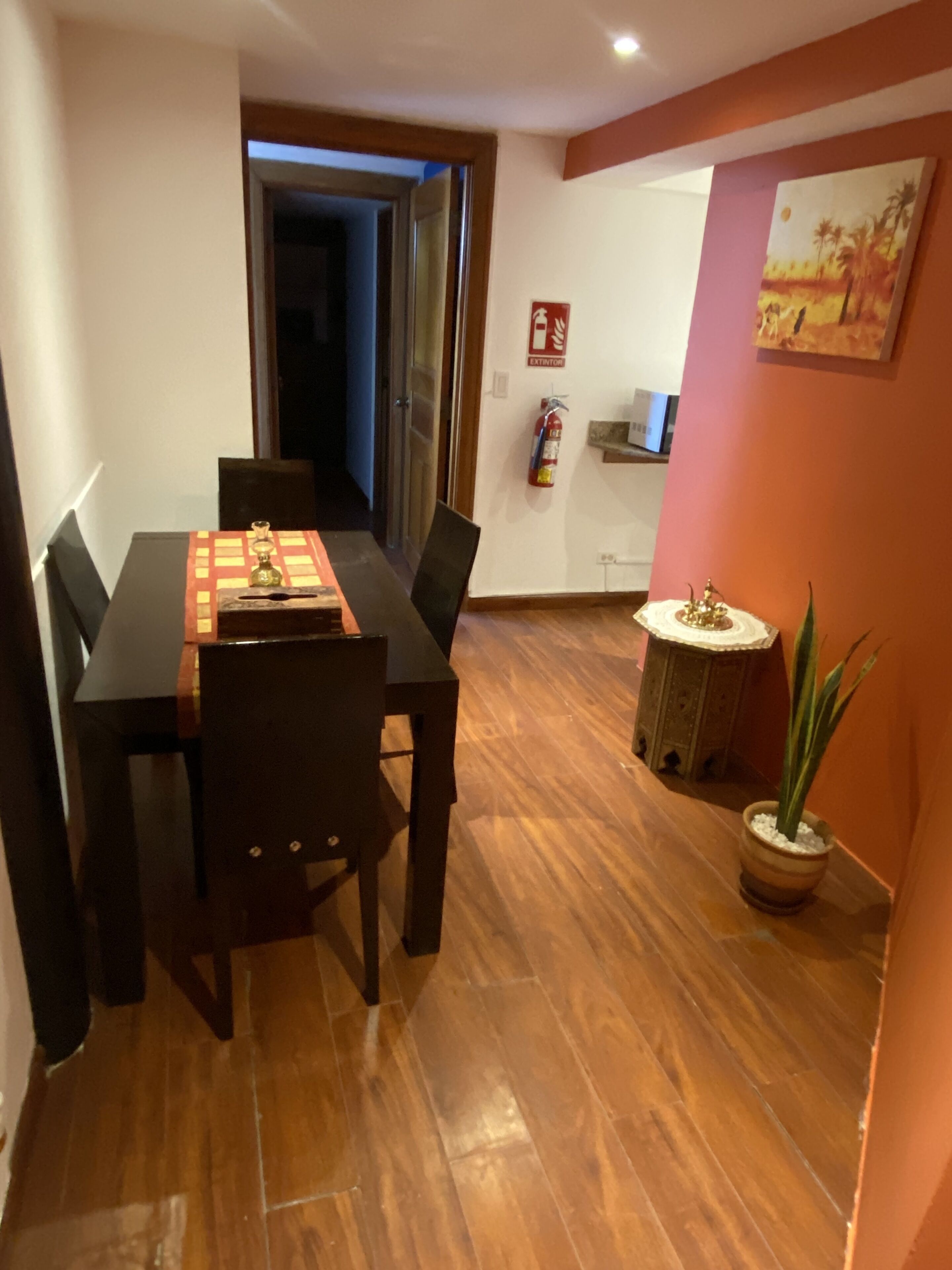 Un coin repas avec une table et des chaises en bois, une petite table d’appoint agrémentée d’un objet décoratif, une plante en pot et un extincteur fixé au mur.