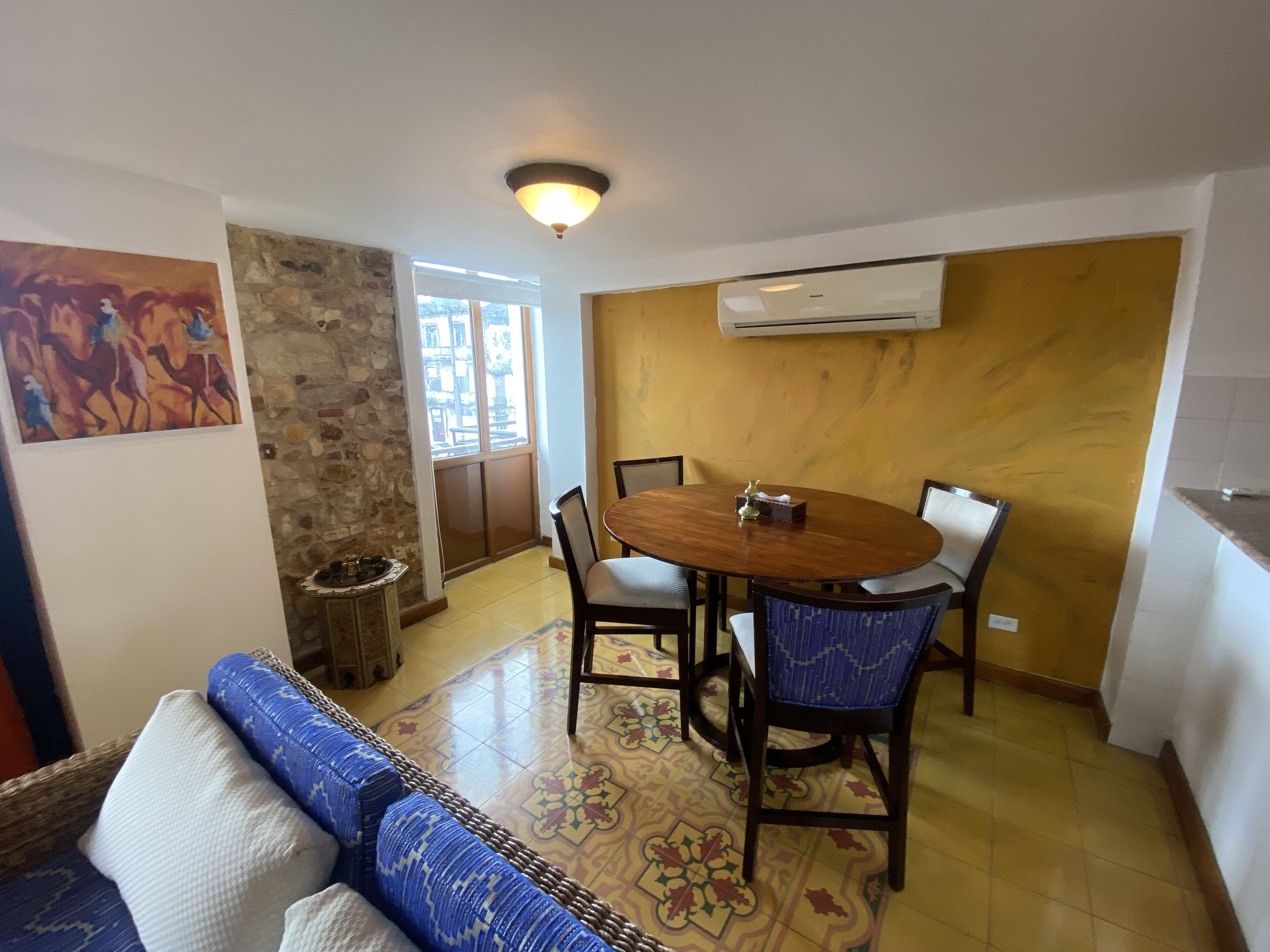 Un comedor con una mesa redonda, sillas y un sofá. Se observa una pared de piedra, un cuadro y un equipo de aire acondicionado en la pared.