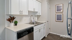 A modern kitchen with white cabinets, a stainless steel sink, and a dishwasher. There are two framed pictures on the wall and a refrigerator to the right. - Kinney #1 xxxxxxxxxx (Austin)