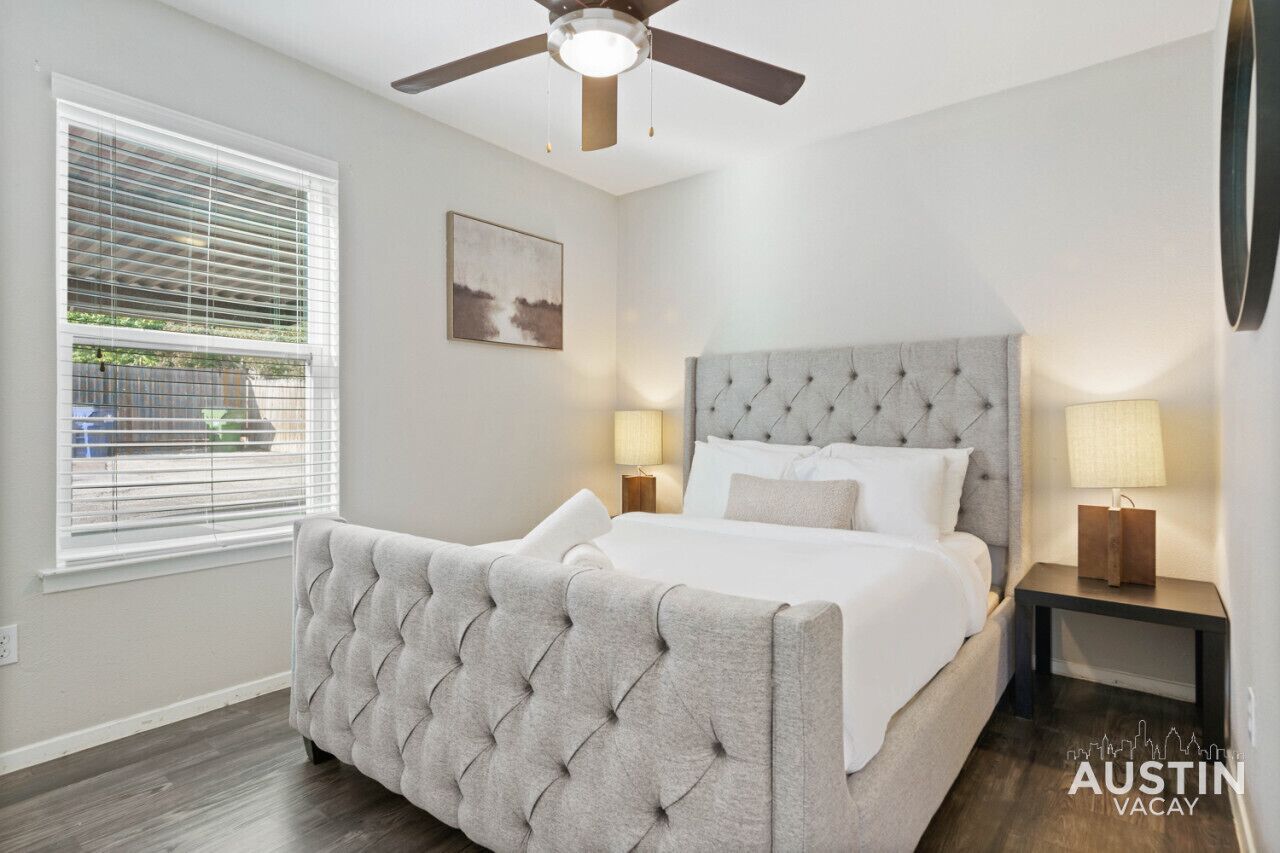 A bedroom with a tufted headboard bed, two bedside lamps, a ceiling fan, and a window with blinds.