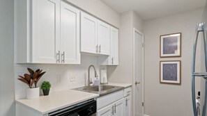 A modern kitchen with white cabinets, a stainless steel sink, and a dishwasher. There are two framed pictures on the wall and a refrigerator to the right. - Kinney #1 xxxxxxxxxx (Austin)
