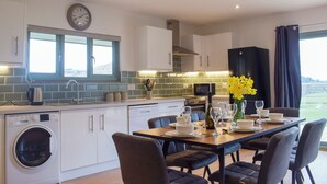 A modern kitchen with a built-in washing machine, a dining area with a table set for four, and a view of the outdoors through a window. - Red Kite Lodge - UK32610 (Stanner, near Kington)