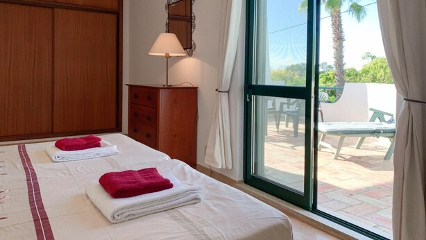 A hotel room with two beds, a wooden wardrobe, a desk with a lamp, and a large window with a view of outdoor furniture. - Casa Josephine by Portucasa (Olhao)