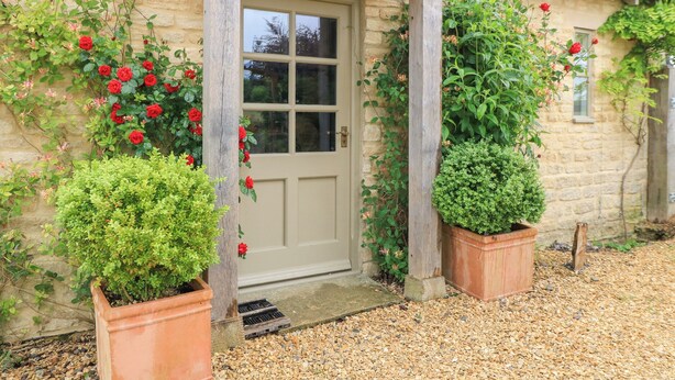 A stone building with a beige door, potted plants, and a gravel driveway. - Pudding Hill Barn Cottage (Cirencester)