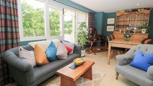 A living room with a grey sofa, a wooden coffee table, and a dining area with a wooden table and chairs. - Pudding Hill Barn Cottage (Cirencester)