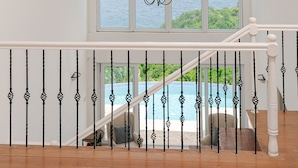 A spacious room with a large window overlooking a pool and greenery, a chandelier, and a staircase with a railing. - Uplifting 3 bed beach island cottage (cap estate)