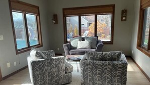 Un salon avec un canapé gris, deux fauteuils, une table basse et un tapis. De grandes fenêtres équipées de stores laissent entrer la lumière, et on y trouve également une cheminée et une vue sur le jardin.