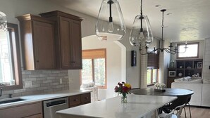 Una cocina moderna con armarios de madera, una isla blanca con taburetes y luces colgantes, y vistas al exterior.
