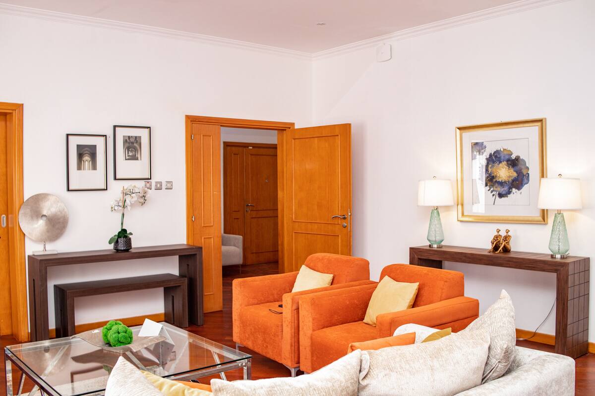 A living room with a glass coffee table, orange sofas, a white armchair, and a wooden console table with lamps.