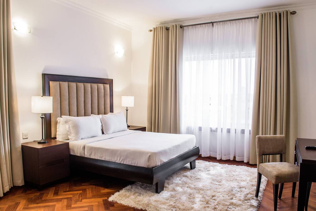 A hotel room with a bed, nightstands, a chair, a desk, and a large window with curtains.