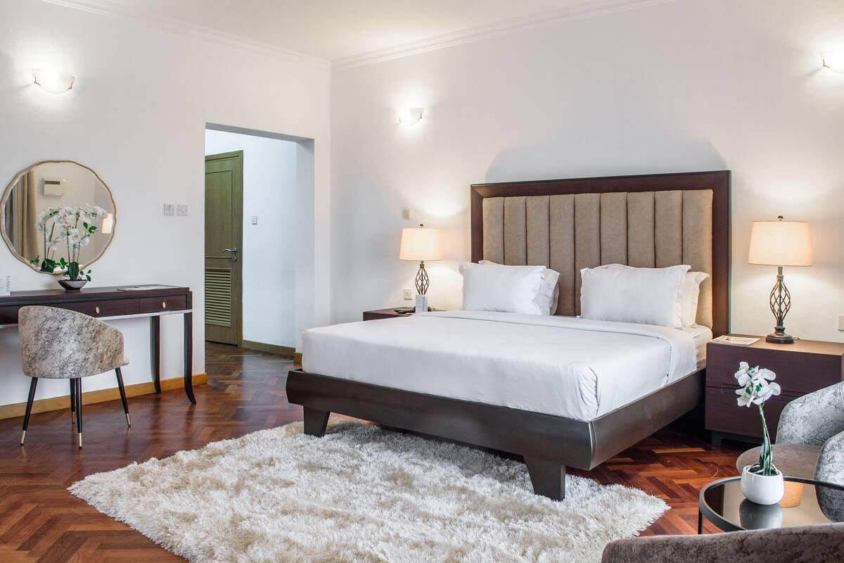 A bedroom with a large bed, two bedside tables with lamps, a desk with a chair, and a mirror on the wall.