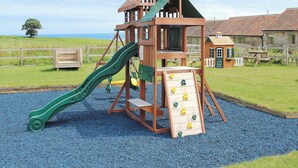 A wooden playground structure with a slide and climbing wall, surrounded by a gravel area and a wooden fence.