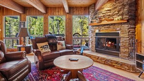Interior acogedor de una cabaña con chimenea de piedra, mobiliario de cuero y grandes ventanales con vistas a un bosque.