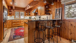 Interior de una cabaña de madera con cocina, barra y ventanas amplias.