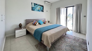 Een moderne slaapkamer met een bed, nachtkastjes, een plafondventilator, een groot raam met gordijnen, en een kleurrijk abstract schilderij aan de muur.