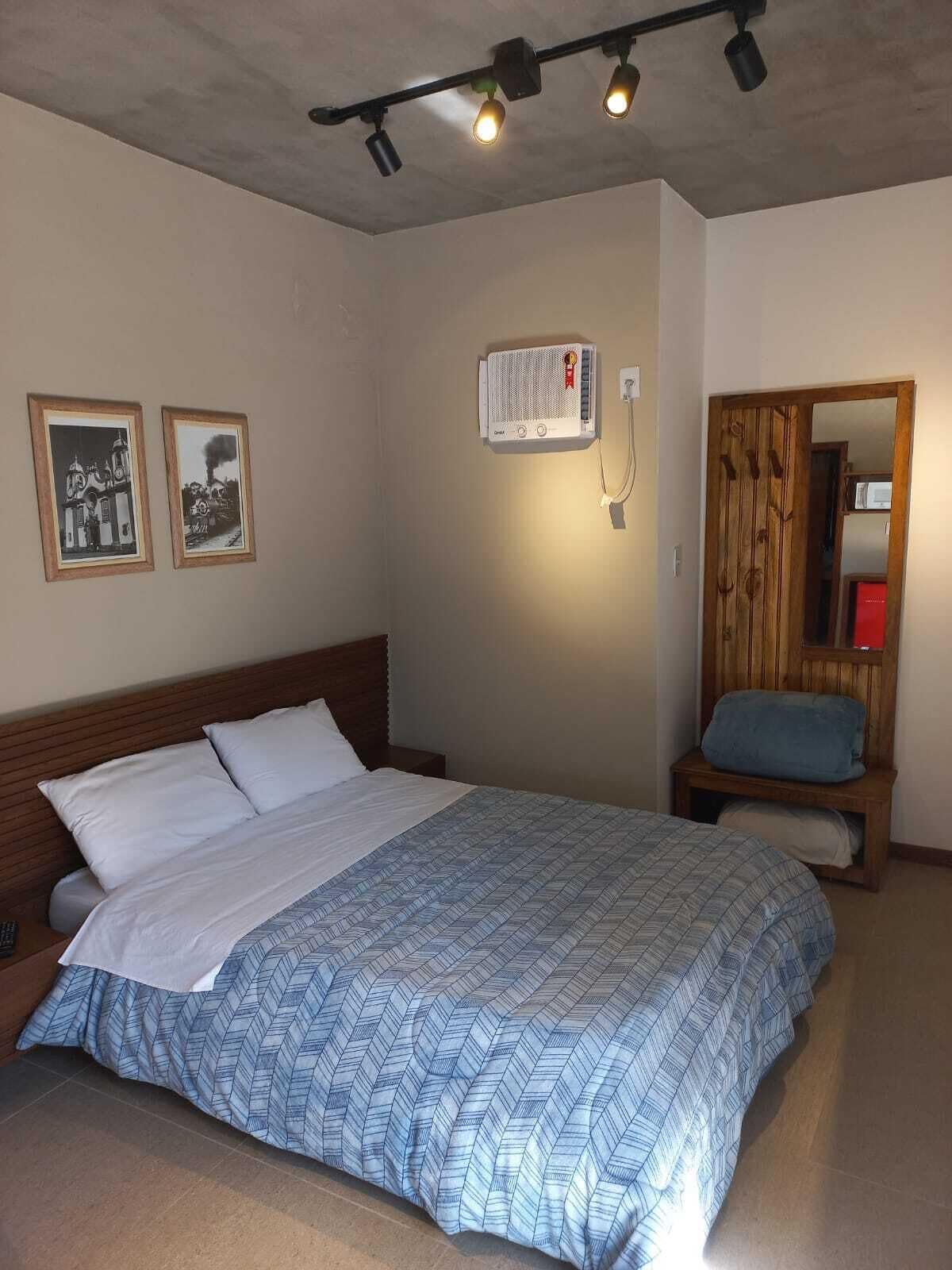 Une chambre à coucher comprenant un lit, une tête de lit en bois, une table de chevet, un climatiseur fixé au mur et deux tableaux encadrés.