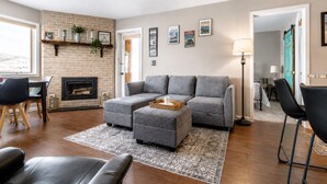 A living room with a fireplace, grey sofa, ottoman, and a coffee table. - Ski In Ski Out Condo in Granby (Granby)