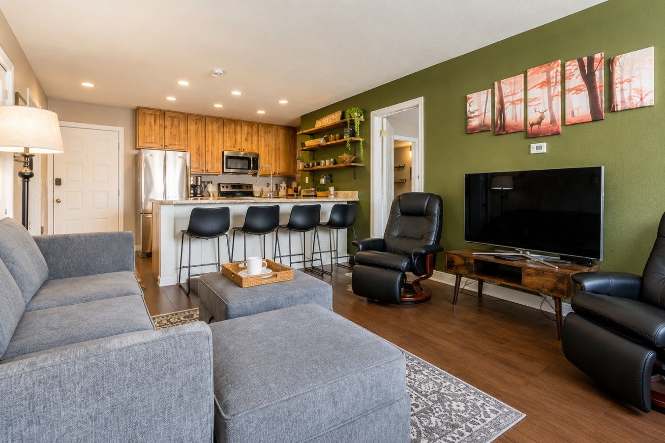 Una sala de estar moderna con un sofá gris, un sillón de cuero negro, un televisor y una cocina al fondo.