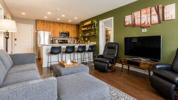 Una sala de estar moderna con un sofá gris, un sillón de cuero negro, un televisor y una cocina al fondo.