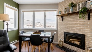 A dining area with a table and chairs, a fireplace, and a view of a snowy landscape through large windows. - Ski In Ski Out Condo in Granby (Granby)