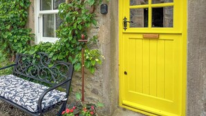 A bench with a patterned cushion, a yellow door, and a potted plant with red flowers. - 1 bedroom accommodation in Linton, near Skipton (Linton, near Skipton)