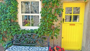 A bench with a patterned cushion, a yellow door, and a window with a view of greenery. - 1 bedroom accommodation in Linton, near Skipton (Linton, near Skipton)