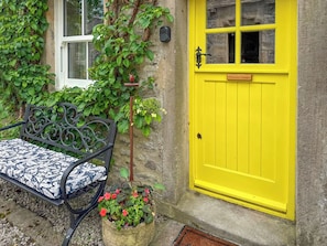 A bench with a patterned cushion, a yellow door, and a potted plant with red flowers. - 1 bedroom accommodation in Linton, near Skipton (Linton, near Skipton)