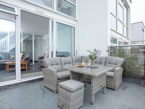 Un espace de détente extérieur moderne, avec un canapé d’angle gris, des fauteuils en osier et une table agrémentée de verres et de fleurs.