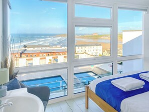A bedroom with a large window, a bed, a bedside table, a sink, and a view of the sea and buildings.