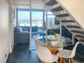 A modern living area with a glass dining table, white chairs, a sofa, and a staircase with stone steps. - 2 bedroom accommodation in Newquay (Newquay)