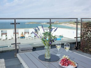 A balcony with a table set for two, featuring a vase of flowers, two glasses of white wine, and a plate of grapes.