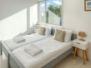 A modern bedroom with two beds, a bedside table, a lamp, and a view of palm trees.