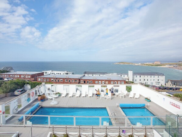 A rooftop pool with two pools, a view of the sea, and a building with a white facade.