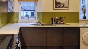 Cocina moderna con estufa integrada, lavaplatos y pileta. Se aprecia una ventana con persianas, un cuadro en la pared y vista a una casa desde afuera.