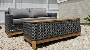 An outdoor seating area with a wicker sofa and a wooden table, set on a gravel surface with a white fence and a brick wall in the background. - Remodeled historic home! Steps away from dining & shops - Easy walk to Old Salem (Winston-Salem)