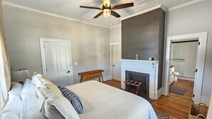 A bedroom with a large bed, a ceiling fan, a fireplace, and a bathroom visible through an open door. - Remodeled historic home! Steps away from dining & shops - Easy walk to Old Salem (Winston-Salem)