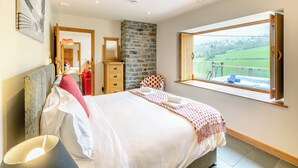 Un dormitorio con cama, cómoda, espejo, pared de piedra y ventana con vistas a un paisaje verde.