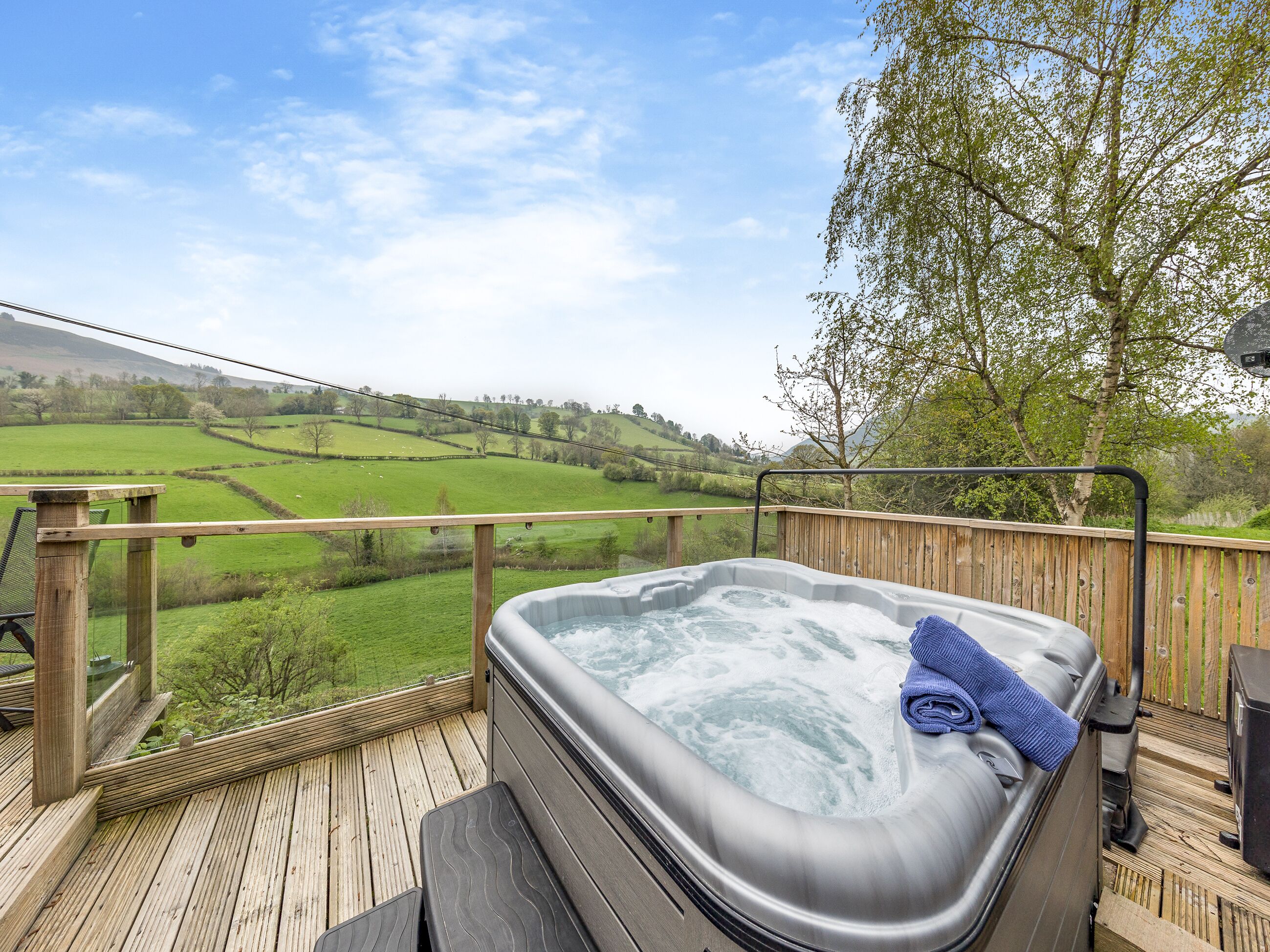 Un bain à remous sur une terrasse en bois, avec vue sur des champs verdoyants et des collines.