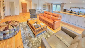 Ein Wohnzimmer mit einer Couch, Sesseln und einem Couchtisch, eine Küche mit Herd und Schränken, Holzboden und ein großes Fenster.
