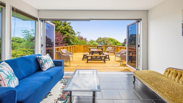 A modern living room with a blue sofa, a yellow bench, and a glass table. Outside, there's a wooden deck with outdoor furniture and a view of greenery.
